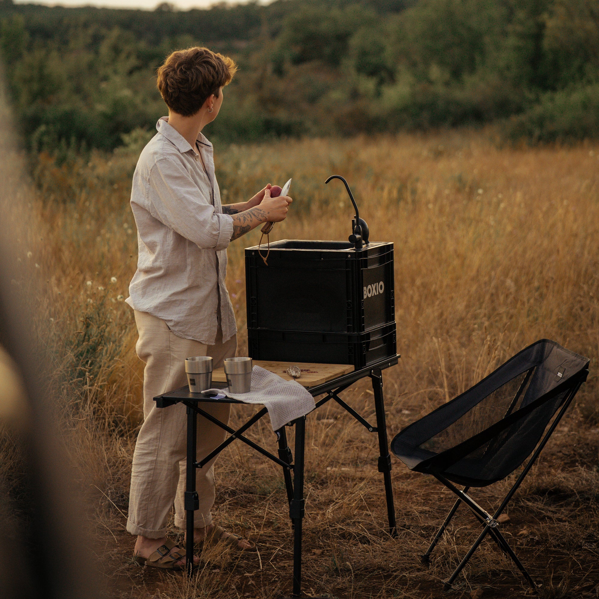 Junge Person mit Tattos steht an einem Tisch im Freien und hält eine Melone, umgeben von hohem Gras.
