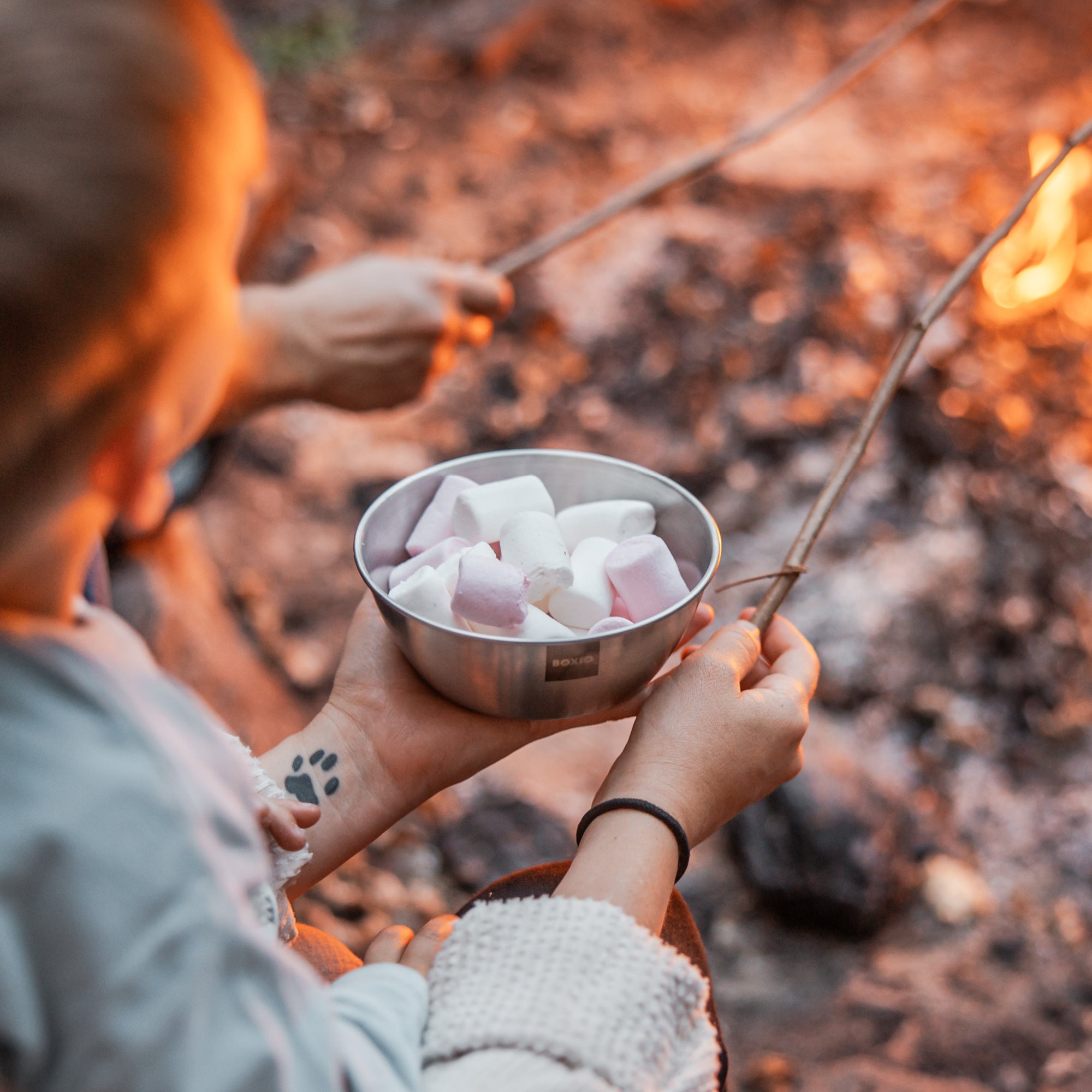 Lagerfeuer im Hintergrund, Frau hält Edelstahlschüssel mit Marshmallows in der Hand und einen Stock