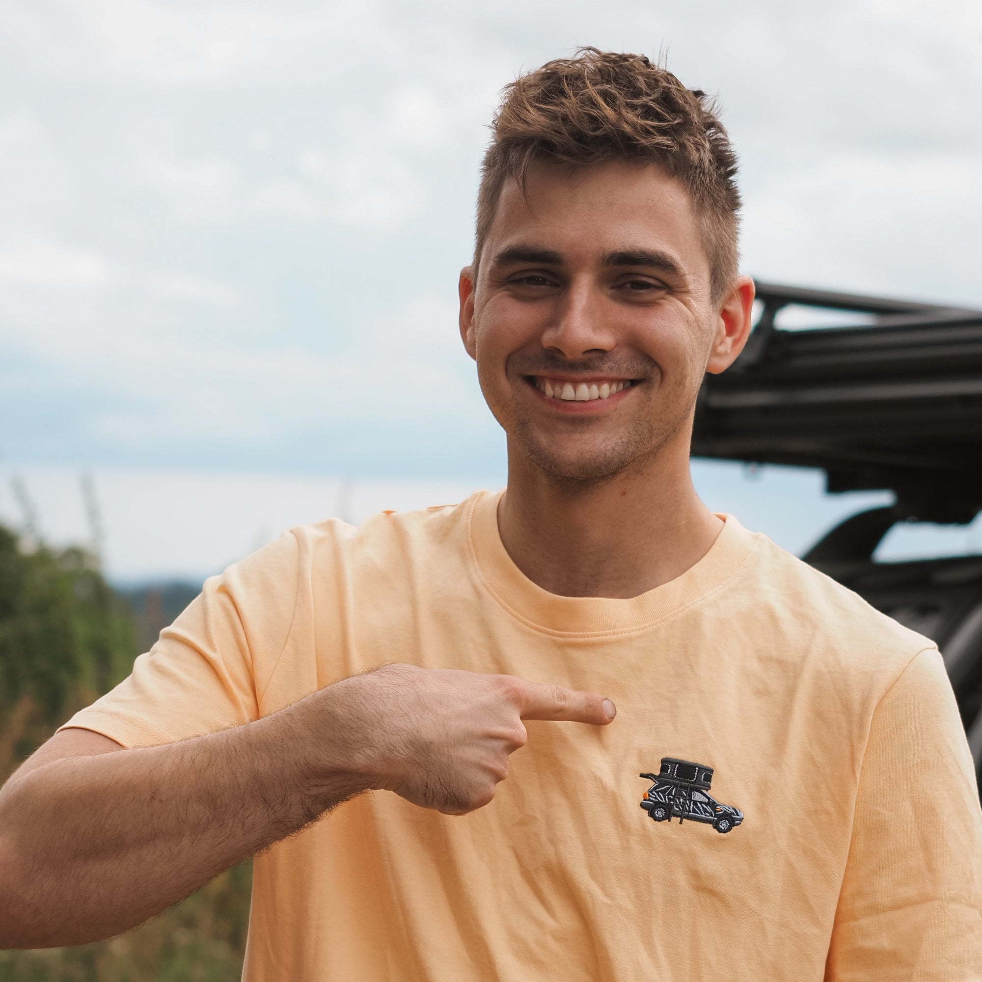 Junger Mann mit kurzem, gestyltem Haar trägt ein gelbes T-Shirt und zeigt lächelnd auf das Auto-Design auf seinem Shirt.