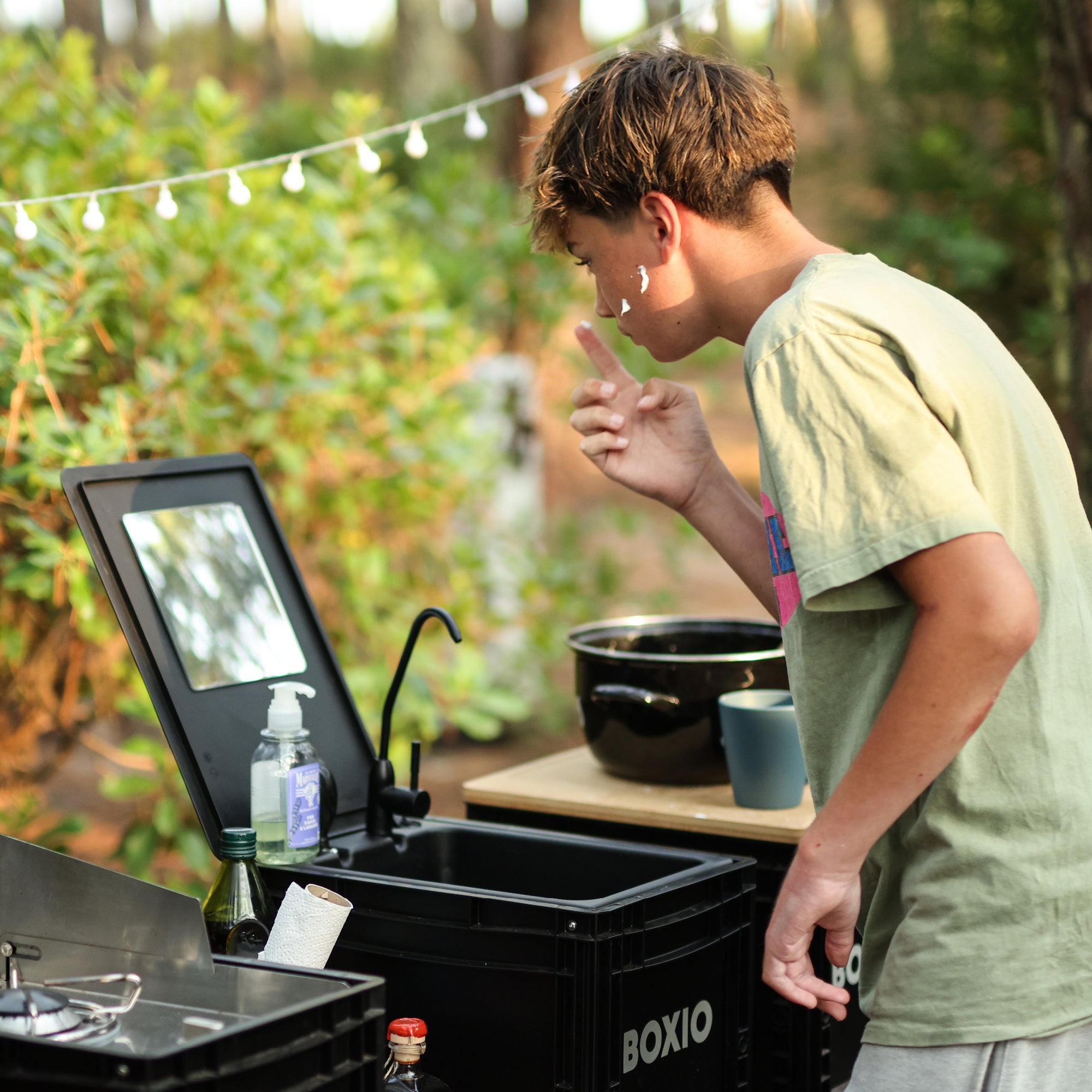 Junger Mann mit Rasierschaum im Gesicht steht vor einem Spiegel in einer naturnahen Campingumgebung.