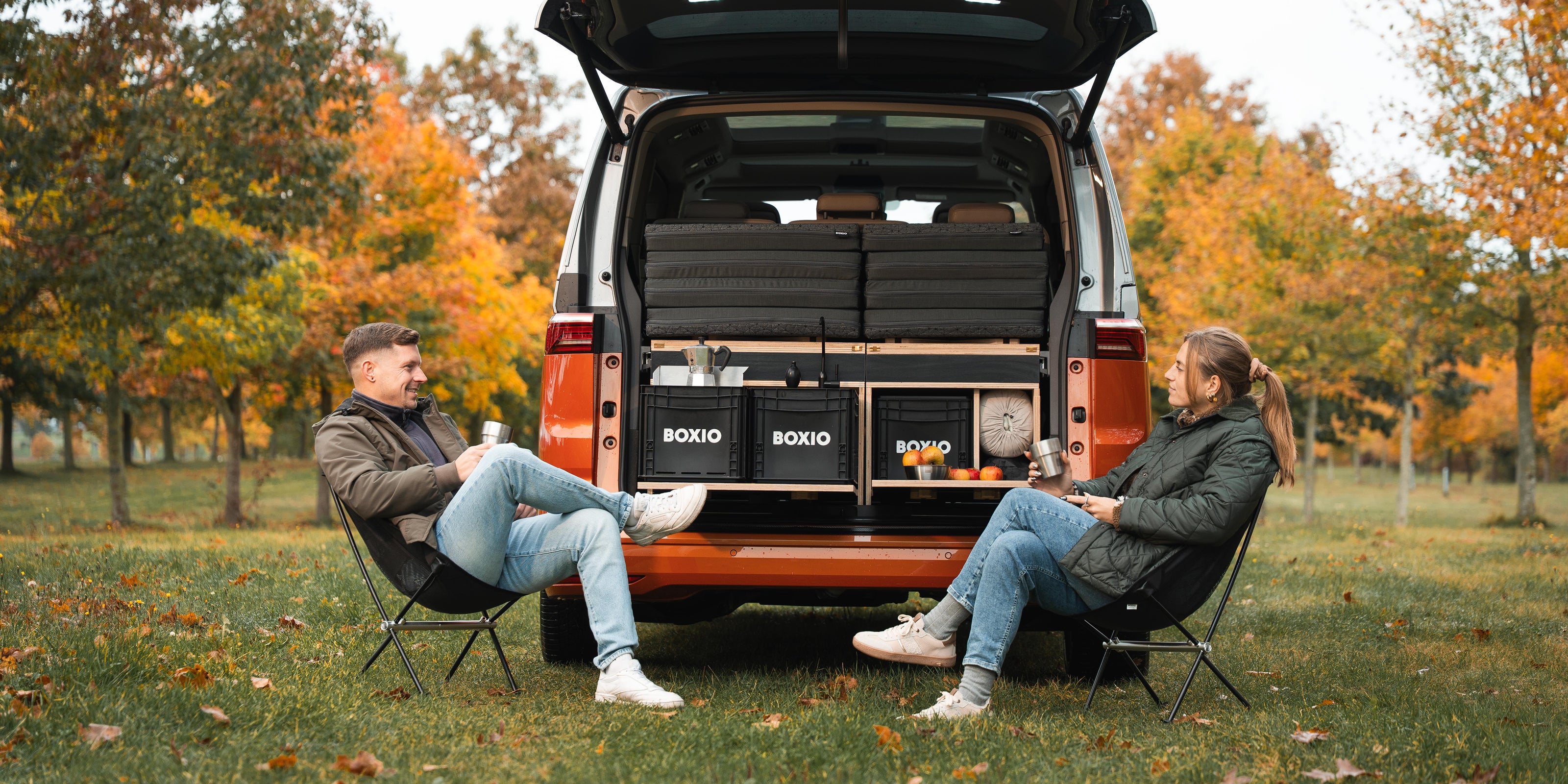 Paar sitzt entspannt vor einem Campingmobil im Herbst, umgeben von bunten Bäumen und Campingzubehör.