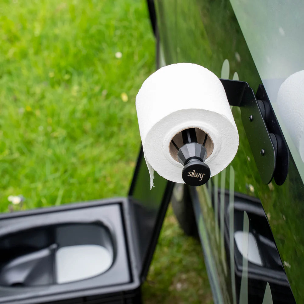 Toilettenpapierhalter mit weißem Papier auf einem schwarzen Träger, im Hintergrund grüne Wiese und eine offene Box.